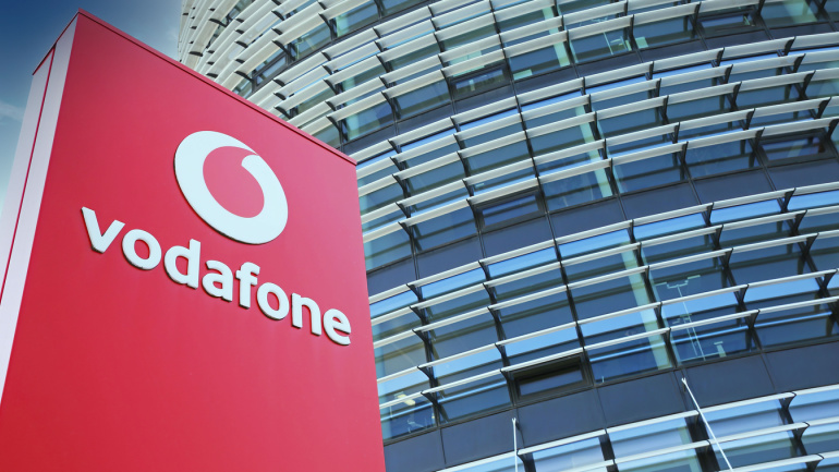 Vodafone in Health. View on red Vodafone logo pillar against futuristic building tower and blue sky, fluffy clouds