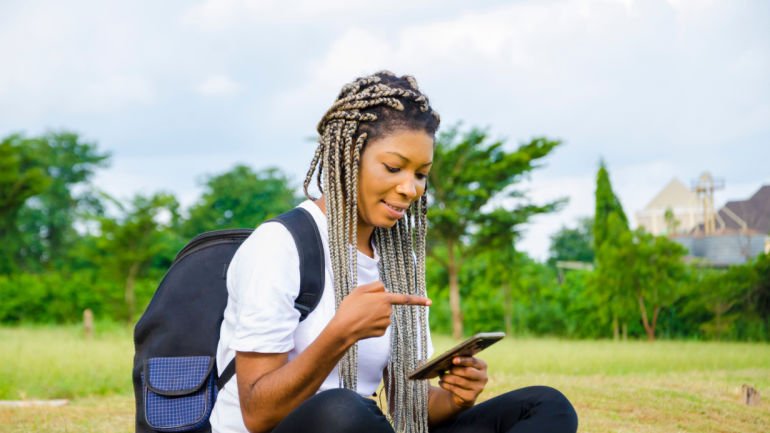 Young beautiful african lady feeling excited as she seats in an open field operating her cellphone, Airtel Nigeria's 5G